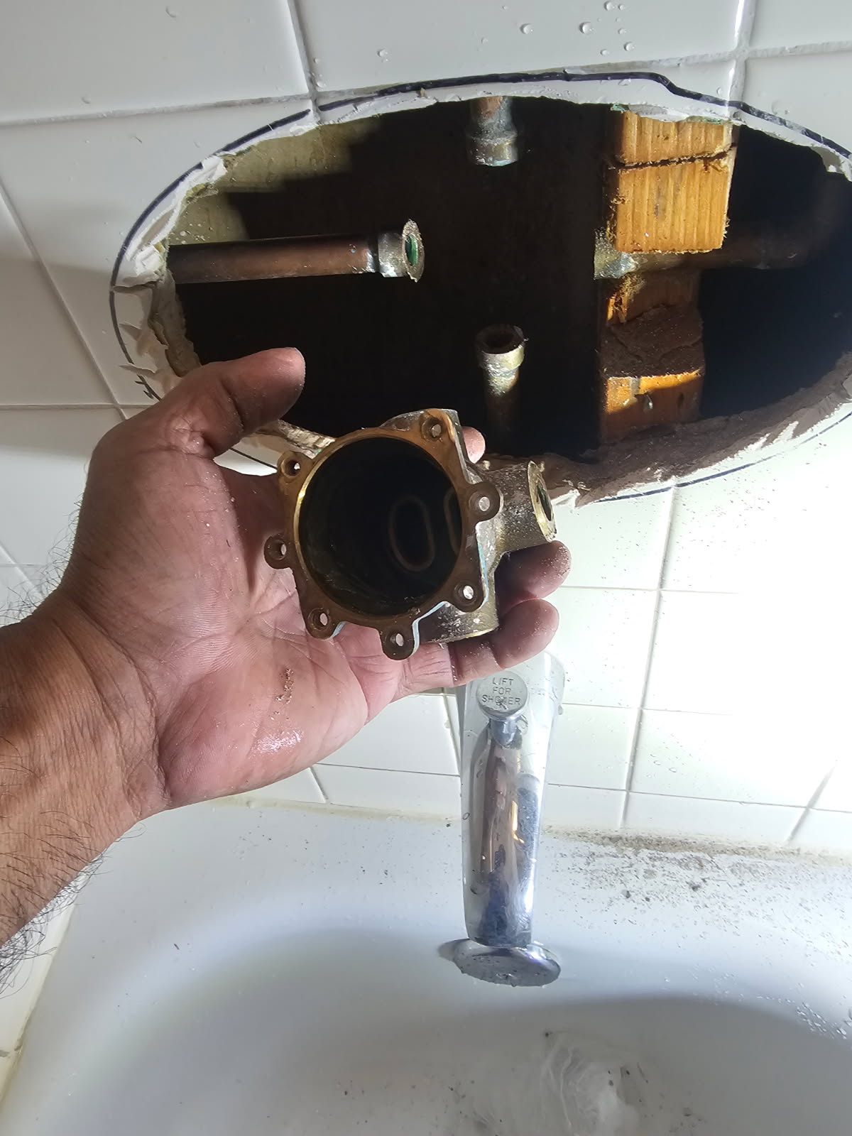 Plumber holding a removed shower valve assembly next to an inspection cut in tile